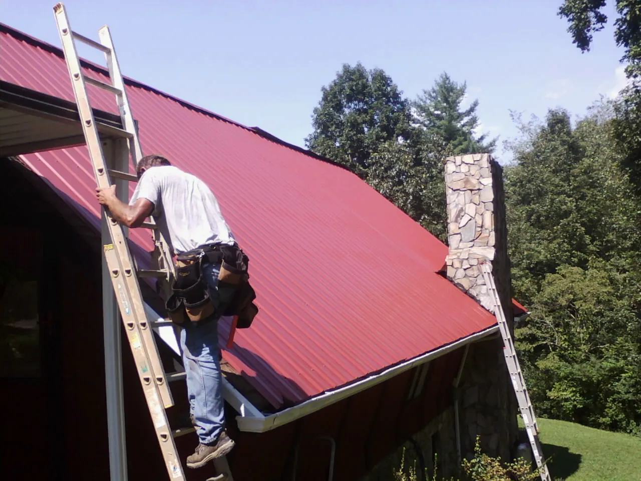 Roofer on a ladder beside a red metal roof and stone chimney — Soffit & Fascia Repair by Roofing Brothers in Oldwick